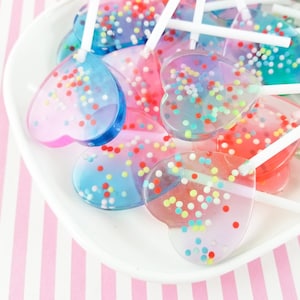 May include: A white plate filled with heart-shaped lollipops. The lollipops are clear and have colorful sprinkles embedded in them. The lollipops are on white sticks.