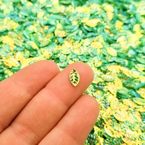 May include: A single gold leaf-shaped sequin with a green center, held in a hand against a background of many similar sequins in green and yellow.
