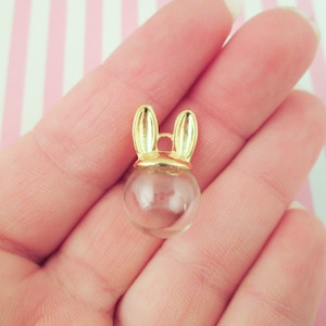 Puede incluir: Un charm dorado con una esfera de vidrio transparente y orejas de conejo. El charm es perfecto para agregar un toque caprichoso a las joyas o las manualidades.