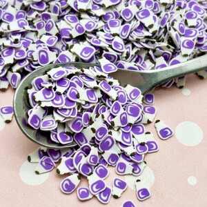 May include: A close-up of a spoon filled with purple and white sprinkles shaped like bottles. The sprinkles are scattered on a pink surface with white polka dots.