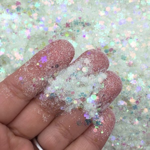 May include: A close-up of a hand holding a handful of iridescent glitter. The glitter is a mix of white, silver, and iridescent colors. The glitter is in a variety of shapes, including circles, stars, and snowflakes.