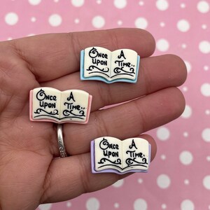 Op de afbeelding: Drie miniatuur open boek bedels met de tekst "Once Upon A Time.." in zwart schrift. De boeken hebben lichtblauwe, roze en lavendelkleurige covers met witte pagina's. De bedels worden in een hand gehouden tegen een roze achtergrond met stippen.