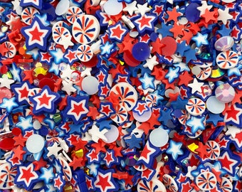 Op de afbeelding: Een close-up van een verscheidenheid aan rode, witte en blauwe sprinkles, waaronder sterren, swirls en cirkels. De sprinkles zijn perfect voor het versieren van cakes, cupcakes en andere desserts.