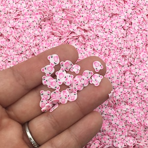 Puede incluir: Una gran cantidad de pequeñas chispas de plástico blancas en forma de cara de cerdo con detalles rosas. Las chispas están esparcidas sobre una superficie y una mano sostiene un pequeño montón de chispas.