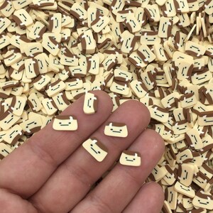 以下が含まれることがあります： 手の中に、小さく黄色と茶色のレジン製のスプリンクルがいくつかある様子のクローズアップ。スプリンクルは、茶色の頭と黄色の底を持つ小さな漫画の顔の形をしています。スプリンクルは表面に散らばっています。