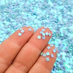 Puede incluir: Una mano sosteniendo purpurina cuadrada azul iridiscente. La purpurina está esparcida sobre una superficie.