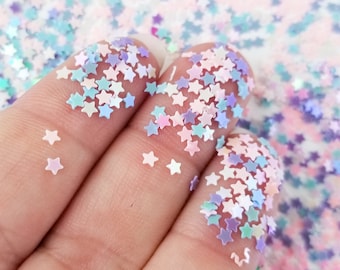 May include: A close-up of iridescent star-shaped glitter in a variety of pastel colors, including pink, blue, and purple. The glitter is scattered on a hand, creating a sparkling effect.