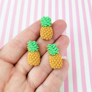 May include: Three small pineapple-shaped charms with green tops and yellow bodies. The charms are on a hand.