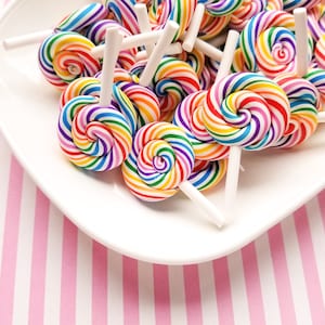 May include: A white plate filled with colorful rainbow swirl lollipops on white sticks. The plate is on a pink and white striped surface.