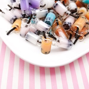 May include: A collection of colorful miniature plastic bubble tea charms with black straws. The charms are in various colors, including pink, blue, purple, and brown. The charms are arranged on a white plate with a pink and white striped background.