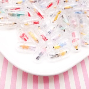 Puede incluir: Una colección de figuras en miniatura dentro de cápsulas transparentes, dispuestas sobre un plato blanco. Las figuras están pintadas en varios colores, incluyendo rojo, amarillo y azul. El plato está sobre una superficie a rayas rosas y blancas.