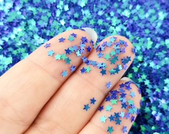 May include: A hand holding blue and green iridescent star-shaped glitter. The glitter is scattered on the fingers and the background is filled with more glitter.
