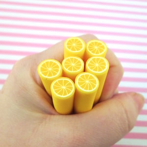 Könnte beinhalten: Eine Ansammlung gelber, zylindrischer Objekte mit orangefarbenen und weißen Zitrusfruchtmustern an den Enden. Die Objekte werden in einer Hand vor einem rosa-weiß gestreiften Hintergrund gehalten. Sie ähneln Bleistiften oder kleinen Dekorationsartikeln.