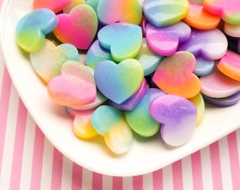 May include: A white plate filled with rainbow heart-shaped resin embellishments. The hearts are iridescent and have a glittery finish.