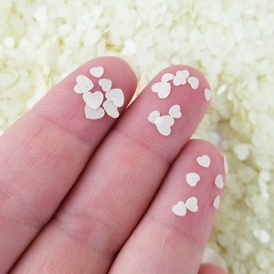 May include: Close-up of small, off-white heart-shaped confetti on fingertips. The confetti is scattered across the fingers and a background of similar confetti. Ideal for crafts, scrapbooking, or party decorations.