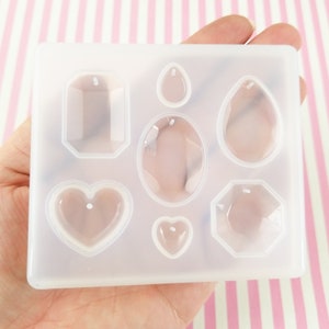 May include: A clear silicone mold with seven different shapes for making jewelry pendants. The shapes include a heart, a teardrop, a rectangle, a rounded rectangle, a hexagon, and two oval shapes.