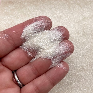 May include: A close-up of a hand holding a pile of iridescent white glitter. The glitter is very fine and has a slight sparkle to it.