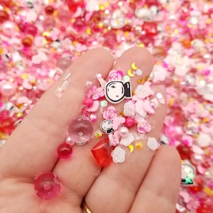 May include: A mix of pink, white, and clear resin charms, including hearts, stars, moons, and a crystal ball. The charms are scattered on a hand.