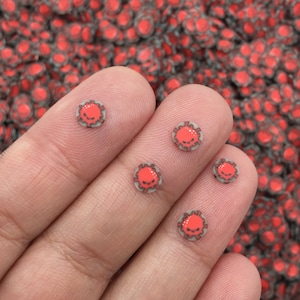 May include: A hand holding five red and gray virus-shaped resin sprinkles. The sprinkles are small and round, and they have a black outline.