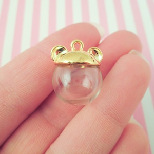 Puede incluir: Encanto dorado con una esfera de vidrio transparente, que presenta una parte superior redondeada y dos pequeños acentos en forma de oreja a los lados.