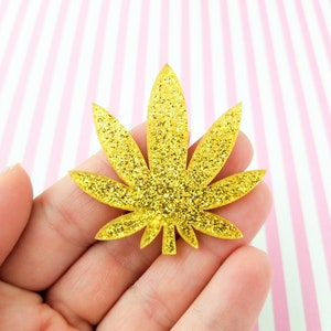 May include: A gold glitter cannabis leaf. The leaf is held in a hand against a pink and white striped background. The leaf has a detailed design with multiple points and a textured, sparkling surface.