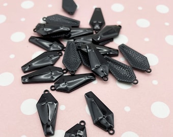May include: A collection of black coffin-shaped charms with small loops for attaching to jewelry. Some charms have a smooth, glossy finish, while others have a textured surface. The charms are scattered on a pink background with white polka dots.