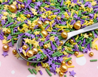 May include: A mix of purple, green, and yellow sprinkles with gold pearl sprinkles. The sprinkles are in a spoon on a pink background with white polka dots.