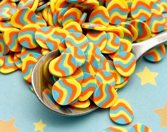 Pode incluir: Um grande monte de granulado amarelo, laranja e azul ondulado em forma oval. O granulado está numa colher de prata sobre um fundo azul claro com estrelas amarelas.