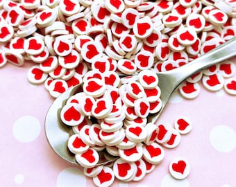 May include: A large pile of white and red heart-shaped sprinkles. The sprinkles are scattered on a pink surface with white polka dots. A silver spoon is partially submerged in the sprinkles.