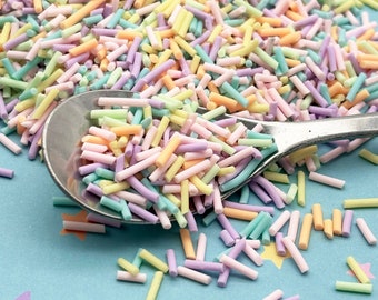 May include: A close-up of a spoon filled with pastel-colored sprinkles. The sprinkles are long and thin, and they are scattered on a light blue surface with yellow stars.