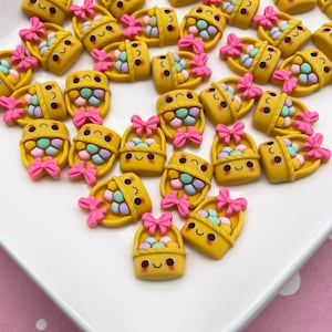 Può includere: Una collezione di ciondoli a forma di cestino pasquale gialli con fiocchi rosa e uova colorate. I ciondoli sono realizzati in pasta polimerica e sono perfetti per aggiungere un tocco primaverile ai tuoi lavoretti.