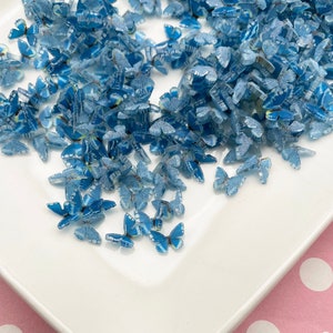 May include: A white plate filled with blue glitter butterfly charms. The plate is on a pink background with white polka dots.