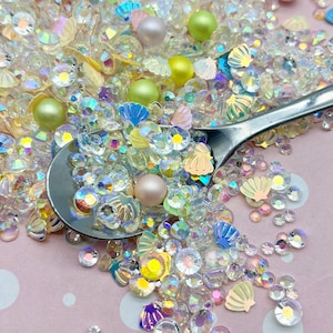 May include: A mix of iridescent clear and pastel-colored glitter, seashells, and pearls. The glitter is in various shapes and sizes, including round, square, and star-shaped. The seashells are in various colors, including white, pink, and blue. The pearls are in various sizes and colors, including white, pink, and green.