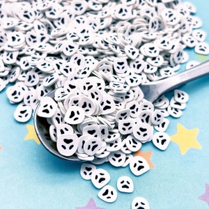 May include: A white spoon filled with white ghost-shaped sprinkles with black eyes. The sprinkles are scattered on a light blue background with yellow and purple stars.