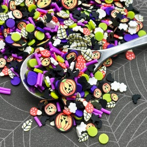 May include: A variety of Halloween-themed sprinkles in a silver spoon. The sprinkles are in various shapes, including pumpkins, stars, leaves, and dots. The colors are black, green, purple, orange, white, and red.
