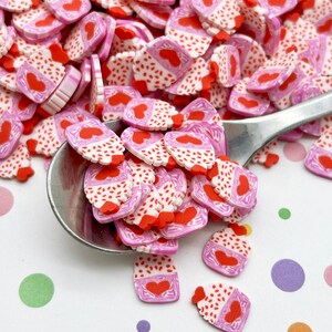 May include: A large pile of pink and white cupcake-shaped polymer clay charms with red hearts and white sprinkles. The charms are scattered on a white surface with colorful polka dots.