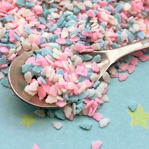 May include: A close-up of a spoon filled with pastel pink, blue, and white sprinkles shaped like hearts and clouds. The sprinkles are scattered on a light blue background with yellow stars.