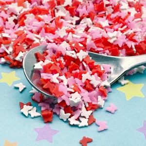 May include: A spoon filled with red, pink, and white butterfly-shaped sprinkles. The sprinkles are scattered on a blue surface with yellow and purple stars.