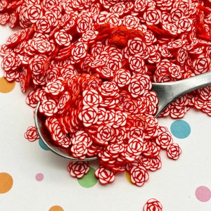 Puede incluir: Un montón grande de rociados de plástico rojo y blanco con forma de rosa. Los rociados están en una cuchara de plata y esparcidos sobre una superficie blanca con lunares de colores.