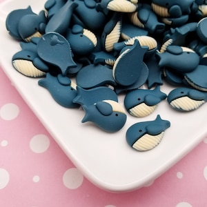 May include: A white plate filled with blue whale-shaped resin charms. The whales have white bellies and are scattered on the plate.