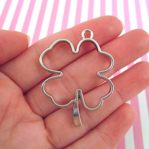 May include: A silver-colored four-leaf clover charm with a loop at the top for hanging. The charm is held in a person's hand against a pink and white striped background. The clover is a symbol of good luck.