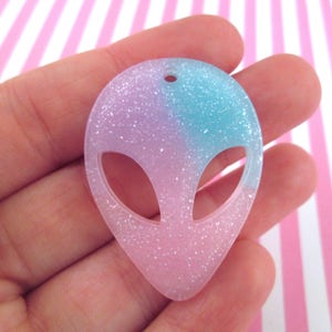 Könnte beinhalten: Ein rosa-blauer Alien-Gesichtsanhänger mit Glitzer, der in das Harz eingebettet ist. Der Anhänger ist in Form eines Alien-Kopfes mit großen Augen und einem kleinen Mund geformt.