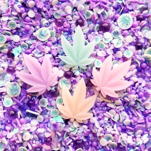 May include: A close-up of a variety of purple, white, and green sprinkles. The sprinkles are in the shape of leaves, stars, and other shapes. There are also some small, round beads in the mix.