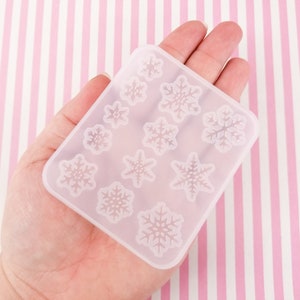 May include: A clear silicone mold with 12 snowflake designs. The mold is held in a hand against a pink and white striped background.