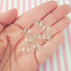 May include: A hand holding several small, clear glass beads. The beads are round with a small hole at the top. The background is a pink and white striped pattern.