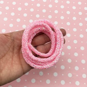 May include: A coiled, pink glitter rope. The rope is a light pink color and covered in sparkling glitter. The background is a light pink color with white polka dots.