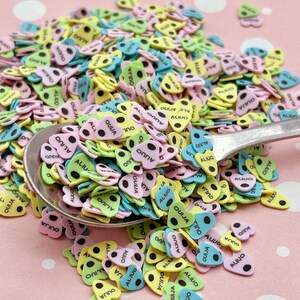 May include: A close-up of a spoon filled with colorful heart-shaped sprinkles. The sprinkles are decorated with black dots and the words "OUIJA" and "ALIUO".