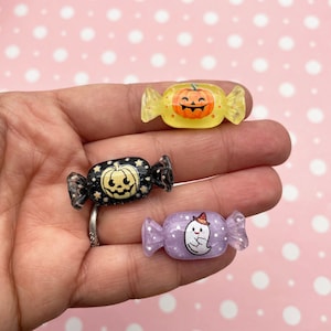 May include: Three Halloween-themed candy charms, each with a different design. One is yellow with a pumpkin face, one is black with a pumpkin face and stars, and one is purple with a ghost wearing a witch hat.