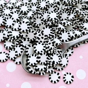May include: A large pile of black and white striped round sprinkles. The sprinkles are arranged in a pile on a pink background with white polka dots. A silver spoon is partially buried in the pile of sprinkles.