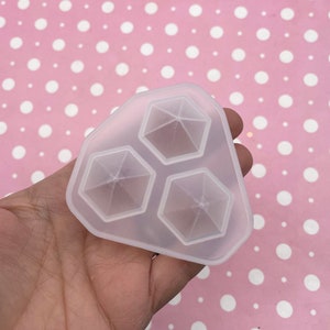 May include: A clear, flexible silicone mold with three hexagonal geometric shapes. The mold is held in a hand against a pink background with white polka dots. The shapes have a starburst design.